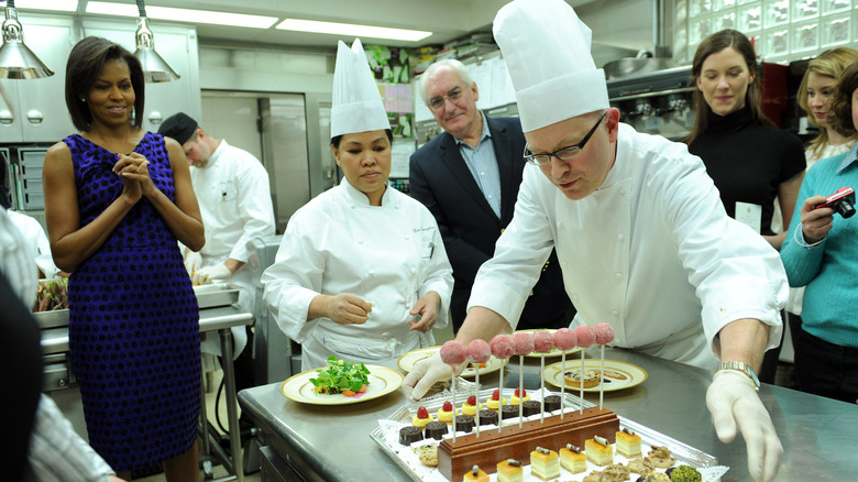 Bill Yosses, Cristeta Comerford, and Michelle Obama standing in the White House kitchen looking at a plate of pastries placed on a metal countertop