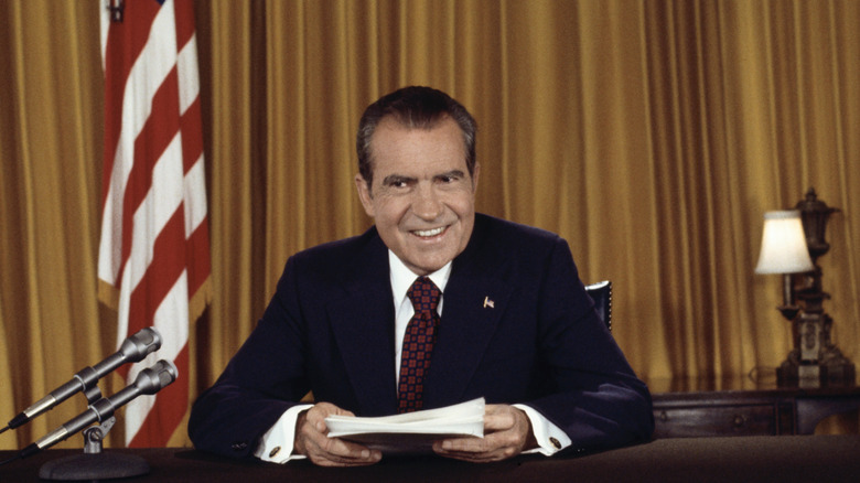 former president Richard Nixon sitting at a desk with papers in hand in front of a gold curtain and two flags