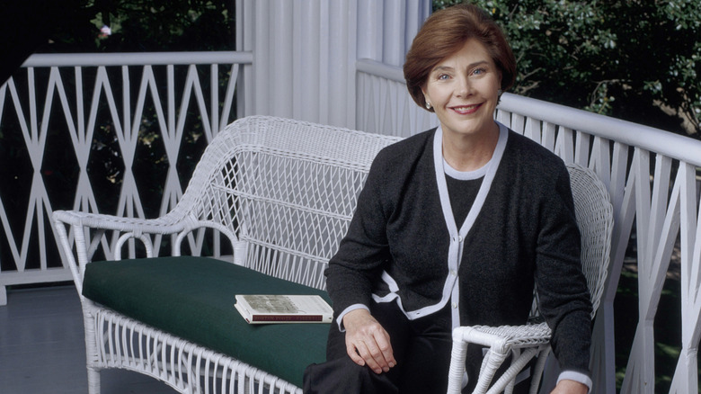 former first lady Laura Bush posing in a black cardigan and blouse while sitting on a white bench
