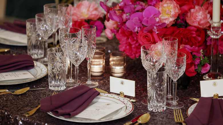 place setting for a Kenyan state dinner with numerous glasses, pink flowers, plates made of china, maroon napkins, and golden utensils placed on the table