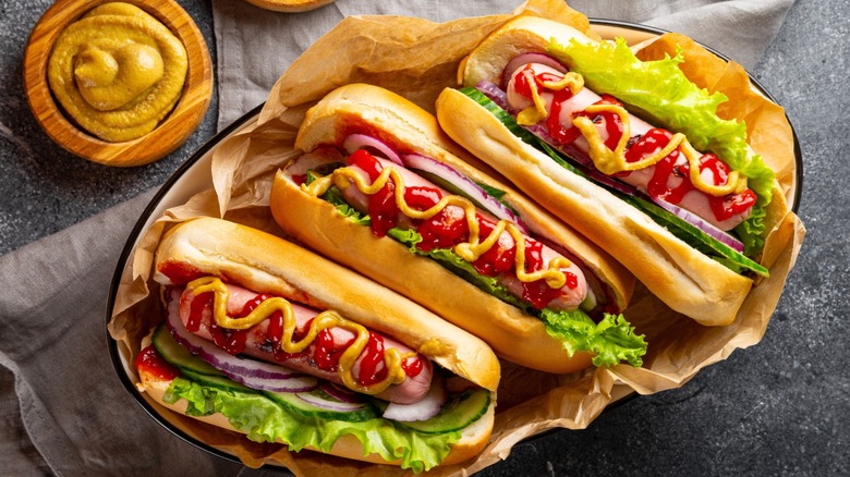 Three hot dogs in buns, topped with ketchup, mustard, red onions, and lettuce, sit on a paper wrapper inside a dish
