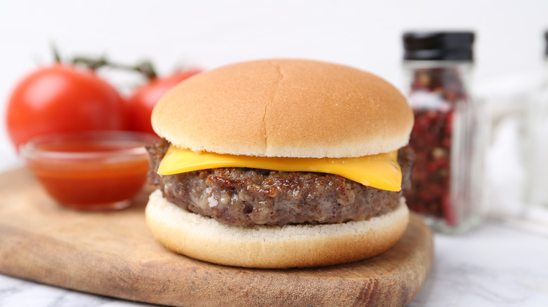 A plain cheeseburger on a wooden cutting board with tomatoes in the background