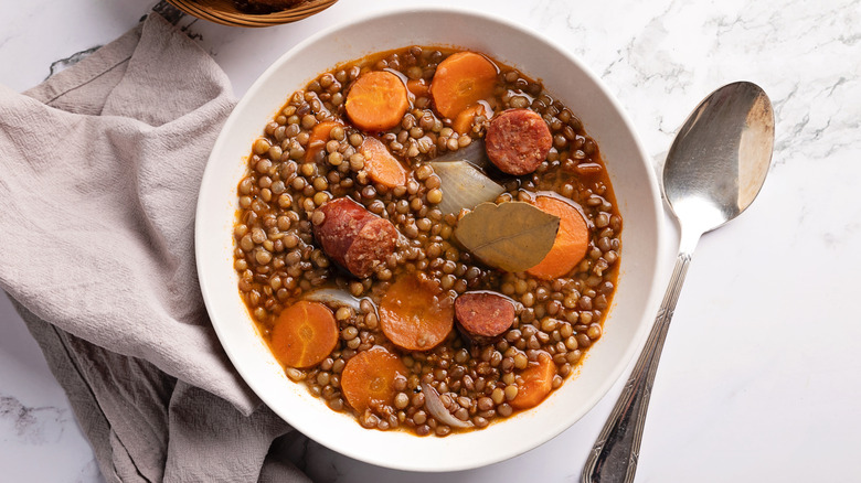 A bowl of sausage, carrots, lentil stew on a white marble table next to a spoon and cloth napkin
