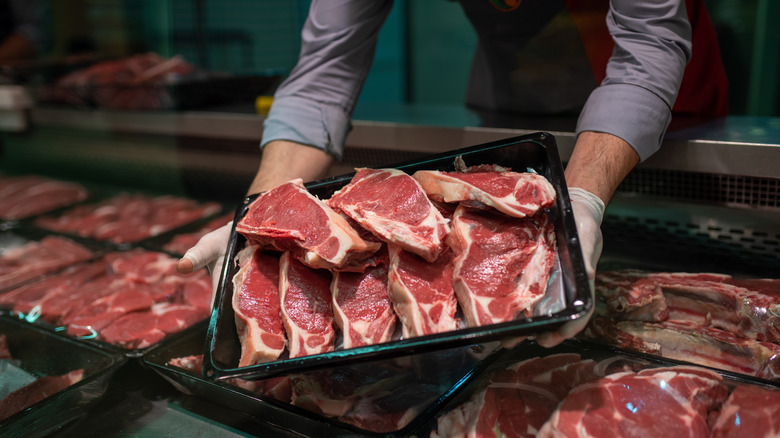 A butcher holds a package of raw bone-in steak cuts over a meat counter