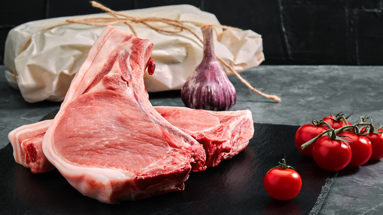 Two raw, bone-in pork chops sit on a black cutting board next to tomatoes, an onion, and a package of meat wrapped in butcher paper