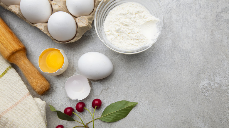 Abowl of flour, eggs (cracked and whole), berries, rolling pin, and dish towel on a gray surface