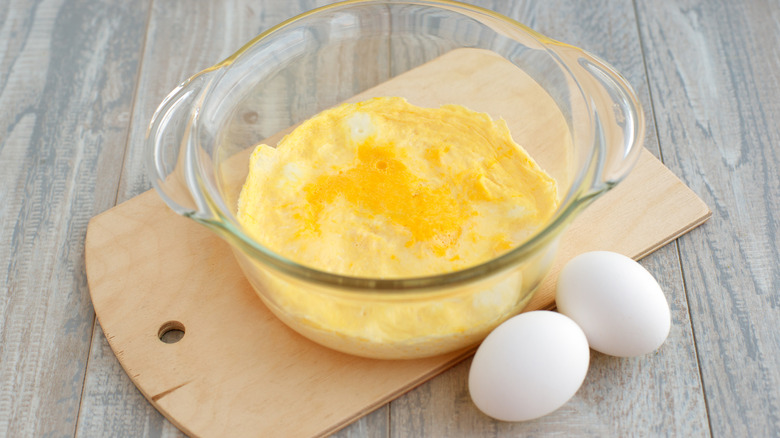 A glass oven bowl filled with cheesy scrambled eggs, on a cutting board, next to two whole eggs.