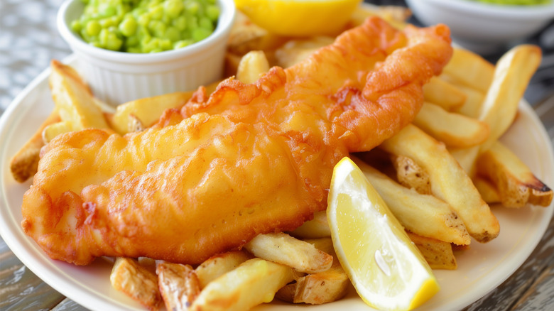 One piece of fried fish on french fries with a small bowl of peas and a lemon wedge on a white plate