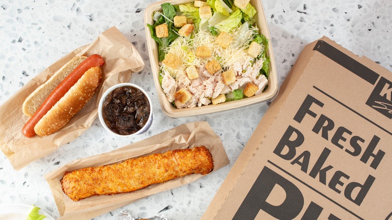 A flatlay spread of Costco pizza food court items, including a salad, hot dog, pizza box, and chicken bake