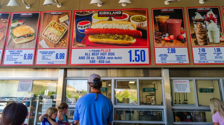 A customer stands in line at the Costco food court, while overhead menu features its hot dog combo, ice cream, and sandwiches