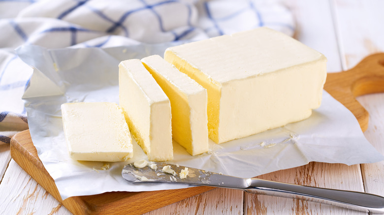 A stick of butter has been cut into with a butterknife, sits in its wrapping on a wooden cutting board