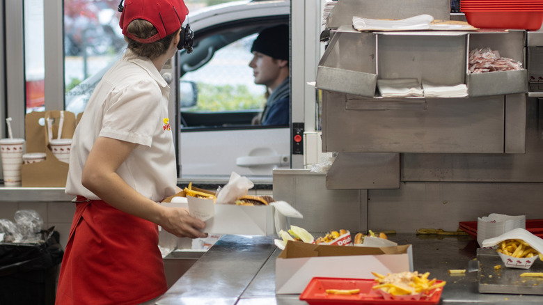 An In-N-Out employee stands at the order window of a drive-thru, building eat-in-the-car boxes.