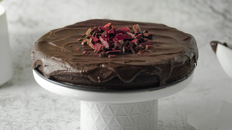 A thin chocolate cake with chocolate frosting sits atop a white cake stand