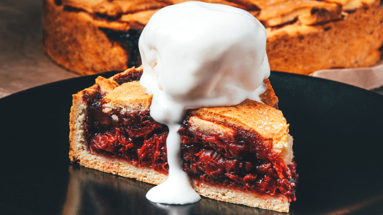 A sice of berry pie on a plate has a round scoop of melting ice cream dripping down the side.