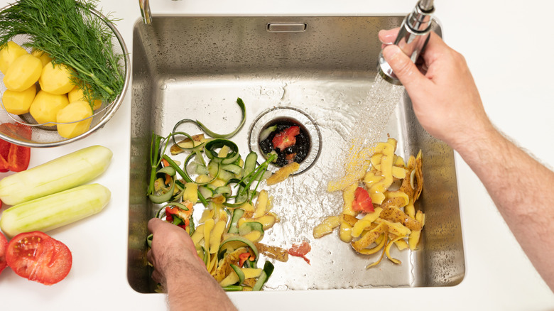 A hand uses a faucet sprayer to wash vegetable peelings down a kitchen sink