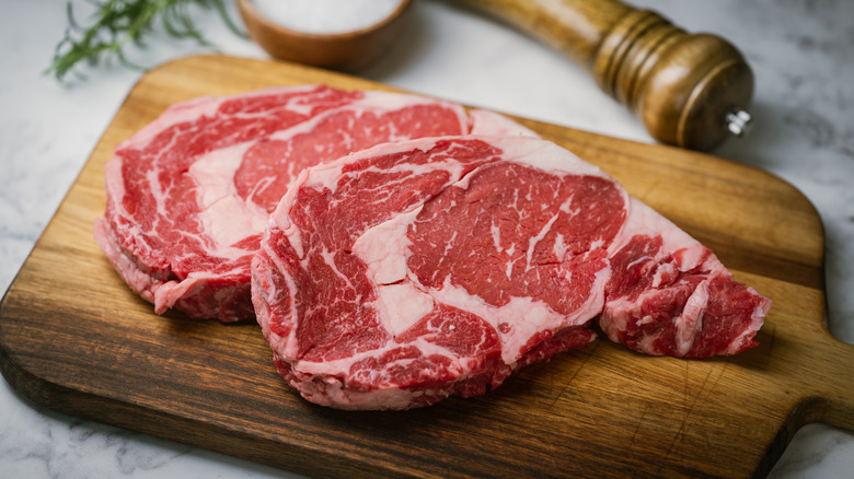 Two ribeyes on a wooden board next to seasonings