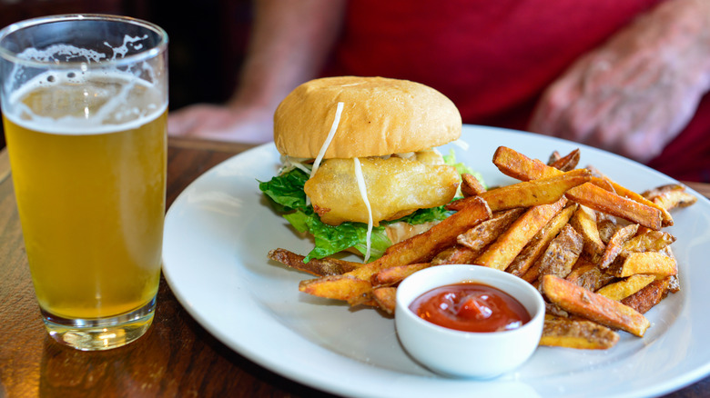 Fried fish sandwich with fries on a white plate with a beer alongside
