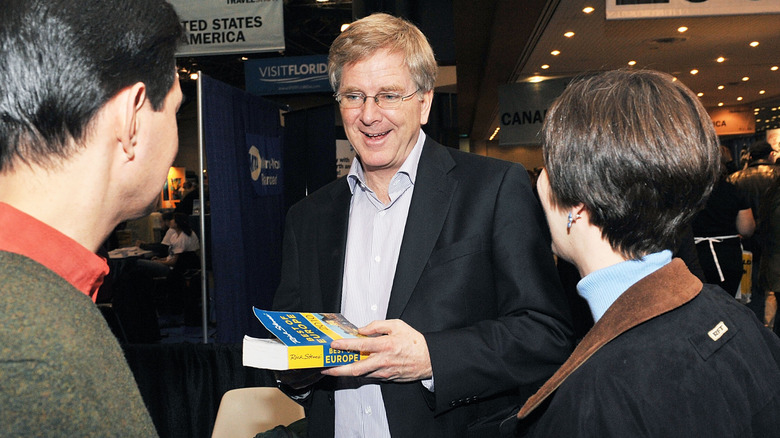 Rick Steves holding one of his Europe guide books and talking to two people
