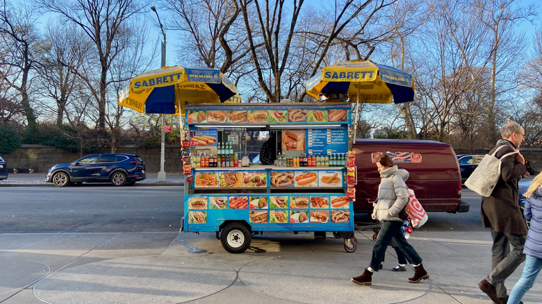 Pedestrians walk by a hot dog cart on an NYC sidewalk