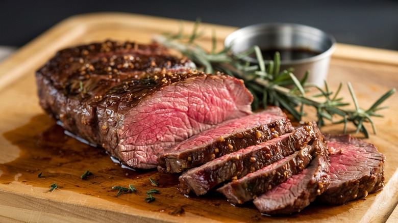 A medium rare jucy beef tenderloin sliced on a cutting board with a sprig of rosemary and a ramekin of dipping sauce in the background.