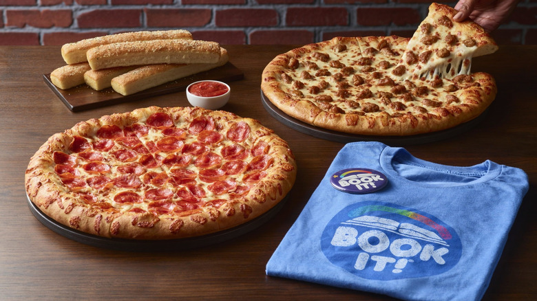 A Pizza Hut pepperoni pizza, sausage pizza, breadsticks, and sauce sit on a wooden surface near a BOOK IT! T-shirt, as a hand removes a slice of sausage pizza from the pan
