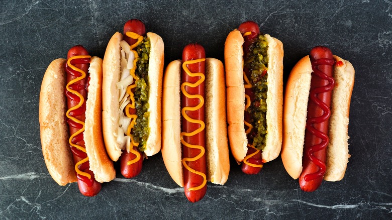Five hot dogs lined up on a stone countertop with various toppings