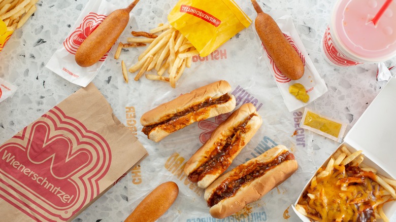 A Wienerschnitzel meal with corn dogs, chili dogs, and fries are spread out on a white granite countertop
