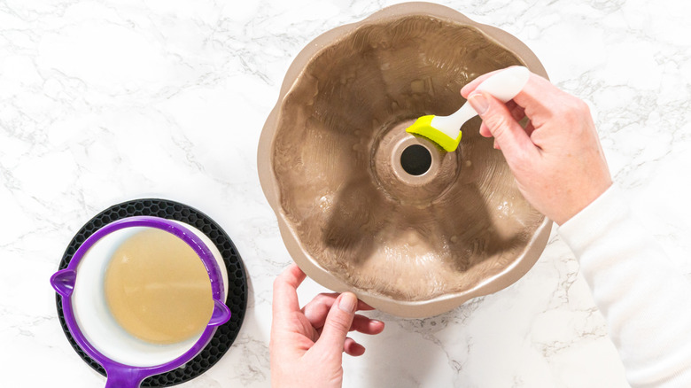 Someone greasing a bundt cake pan with a butter brush on a white marble table