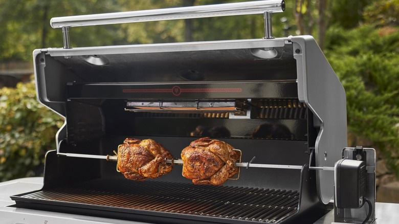 Two chickens turn on a spit over a gas grill.