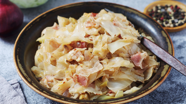 Fried cabbage with some sliced bacon in a black bowl, with an onion and a bowl of lemon peppercorns in the background.