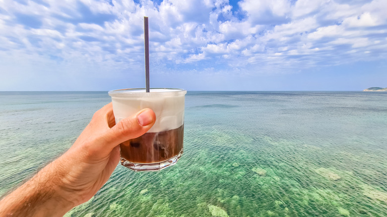A small iced latte with dense, creamy milk held in front of a vast oceanic scene.