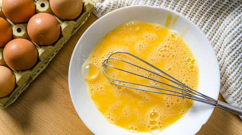 An egg mixture and metal whisk sit in a white bowl, with a cart.on of eggs sitting adjacent