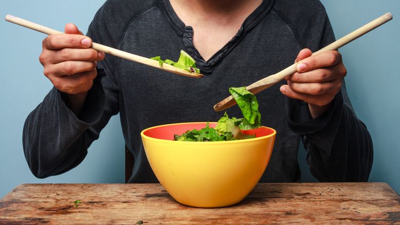 Man tossing salad greens with wooden spoons in a large yellow bowl