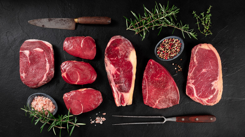 Various cuts of beef on a black surface with herbs and spices