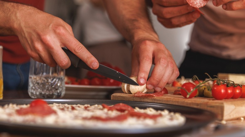 Closeup on the hands of two people as they place toppings on a pizza.