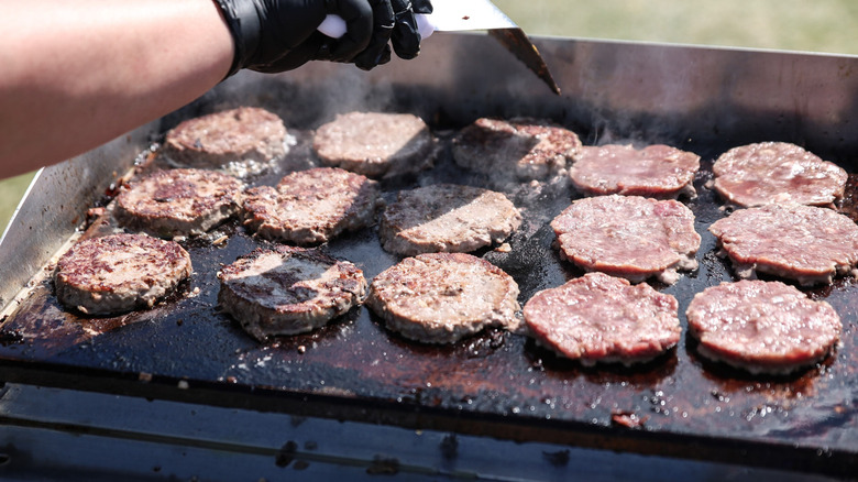 A person with a gloved hand uses a turner to flip burgers on a flat top grill