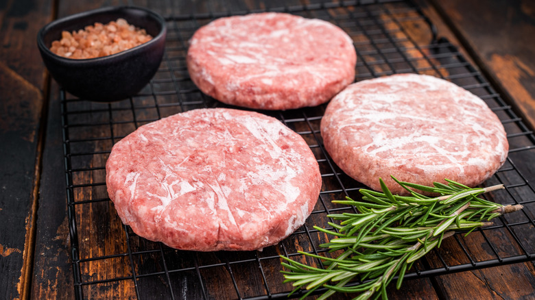 Frozen beef patties on a cooling rack set atop a wood board with salt and rosemary