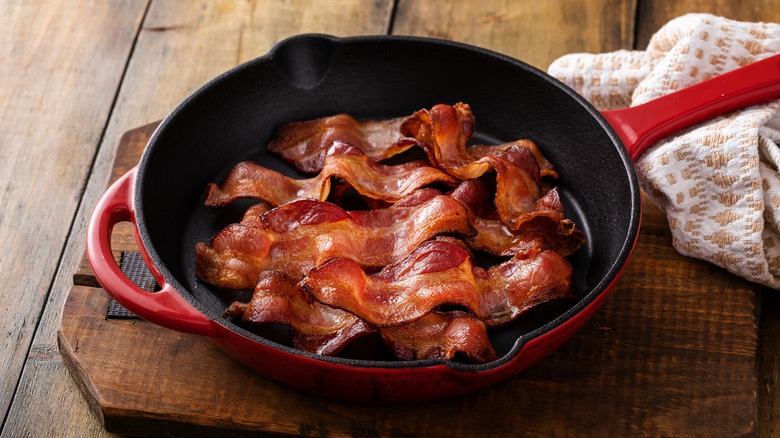 cooked bacon in a skillet with red handles on a wooden cutting board on a wooden table