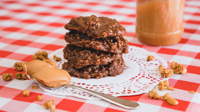 A stack of no-bake peanut butter chocolate cookies sit on a doily, under a red and white checkered tablecloth next to a scoop of peanut butter on a spoon