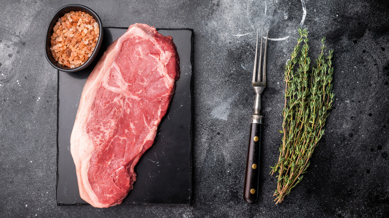 A strip steak next to herbs, salt, and a fork