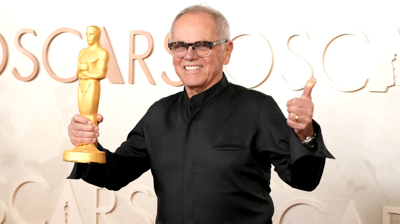 Wolfgang Puck in a black shirt holding an Oscar award in front of  an Oscars promotional background