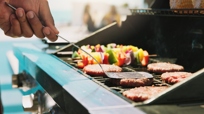 A hand presses a burger patty on a crowded grill alongside vegetable skewers.