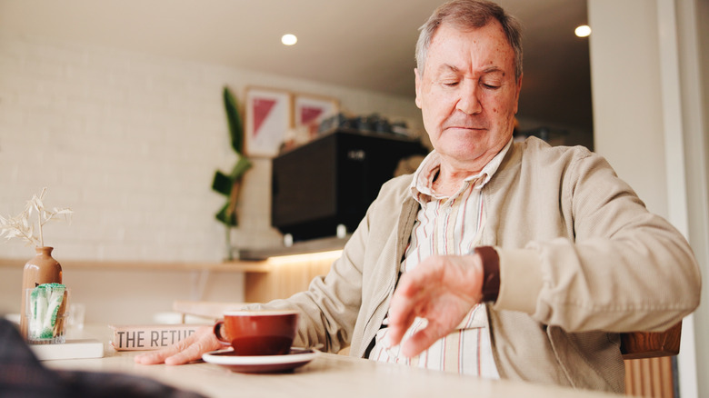 Disgruntled senior looking at a watch while having a cup of copy.