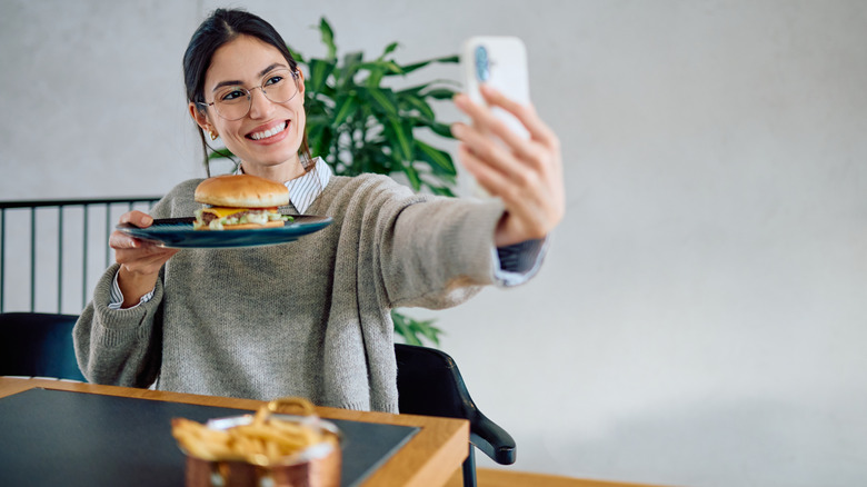 A person smiling while taking a selfie with a hamburger.