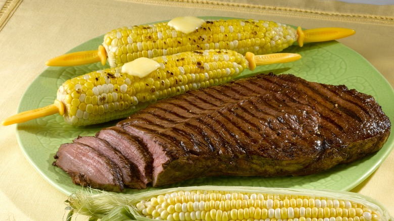 Grilled London Broil resting on a plate beside grilled corn