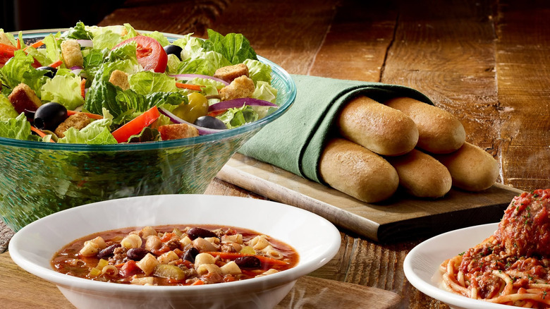 A salad from Olive Garden sits next to breadsticks, soup, and pasta.