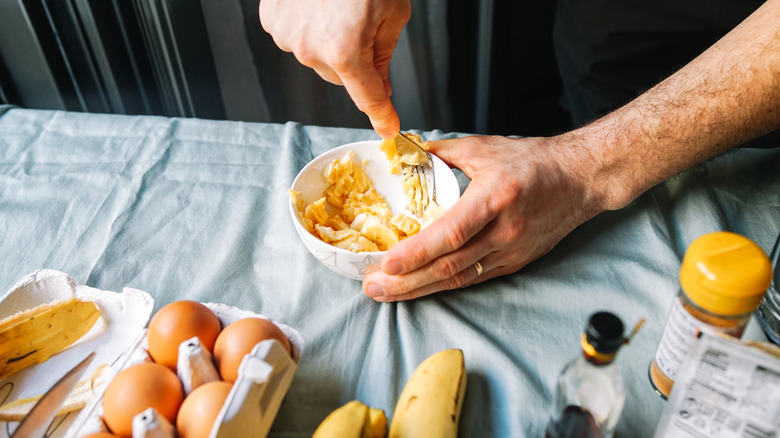 A person mashes bananas and eggs with a fork in a small bowl