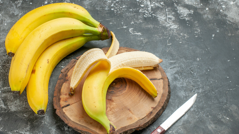 A bunch of yellow bananas next to a partially peeled banana on a wooden cutting board