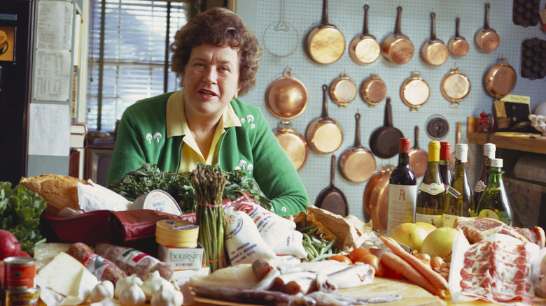 Julia Child in her kitchen with a table full of produce, bread, cheese, cured meats, and wine.