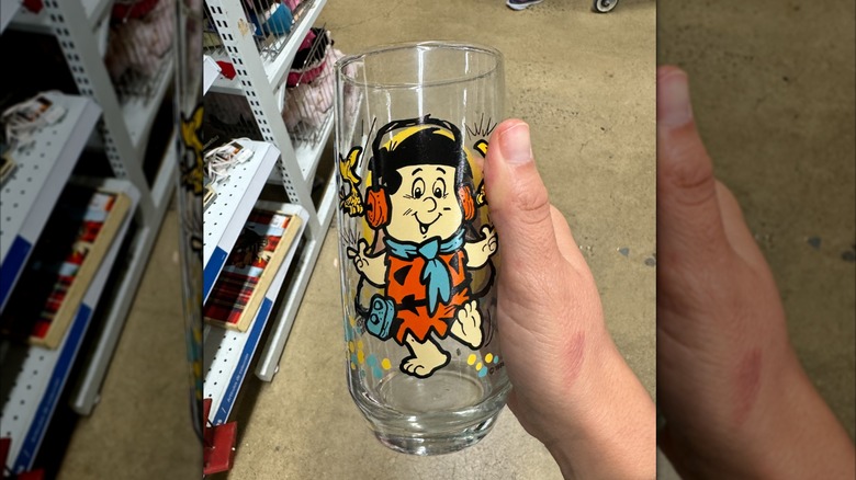 A hand holds a promotional drinking glass featuring an image of young Fred Flintstone